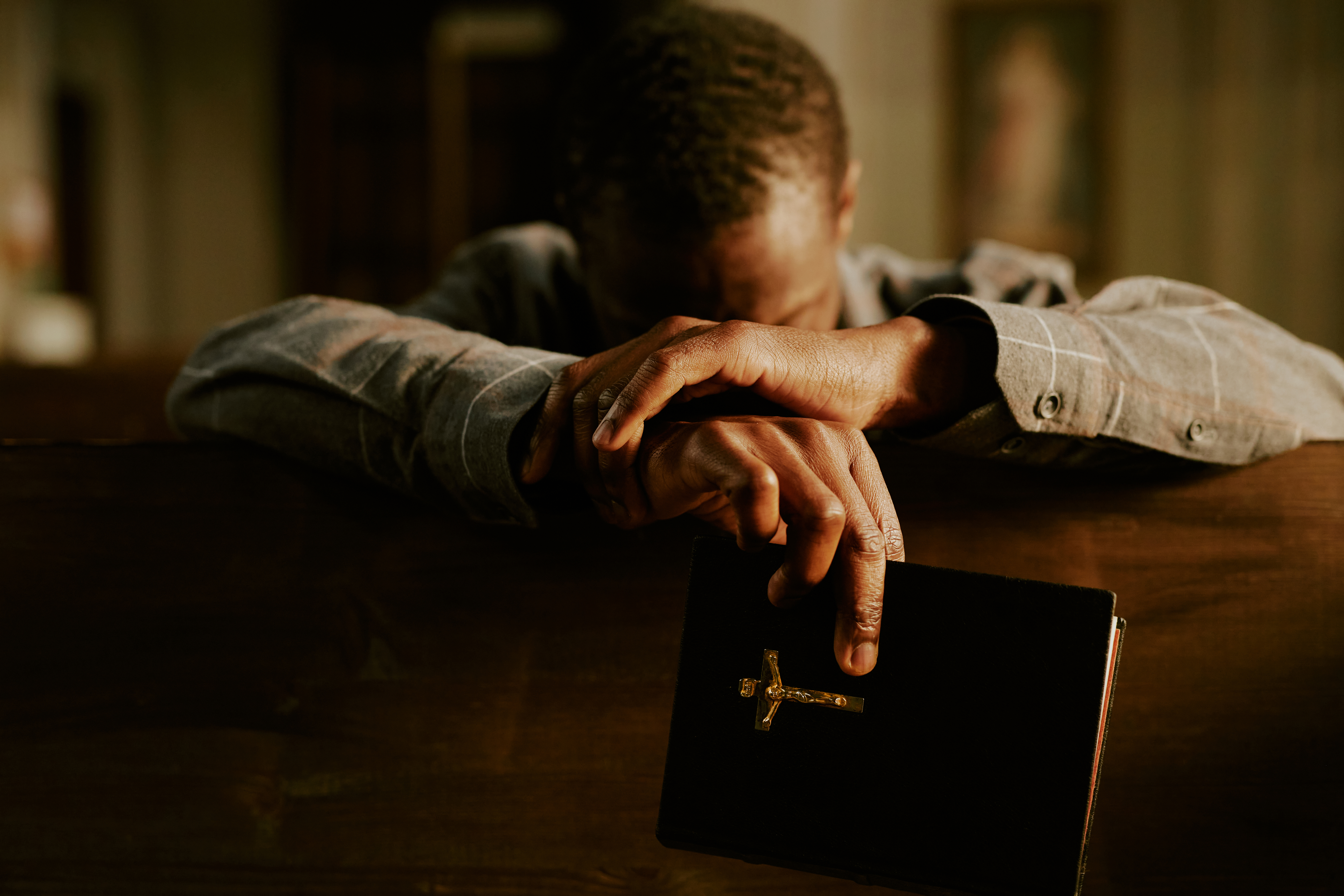 Medium closeup of unrecognizable Black man sitting in Catholic church with Bible in hand praying to God